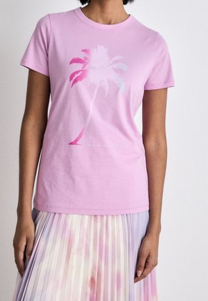 Vrouw draagt een lichtroze T-shirt met korte mouwen en een vervaagde afbeelding van een palmboom, gecombineerd met een geplooide rok in pastelkleuren.