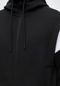 Μαύρο φούτερ με φερμουάρ και κορδόνια, με ανάγλυφο λογότυπο "BOSS" στο στήθος, υφασμένο από υφή υφάσματος και διακριτική λευκή λεπτομέρεια στο μανίκι.