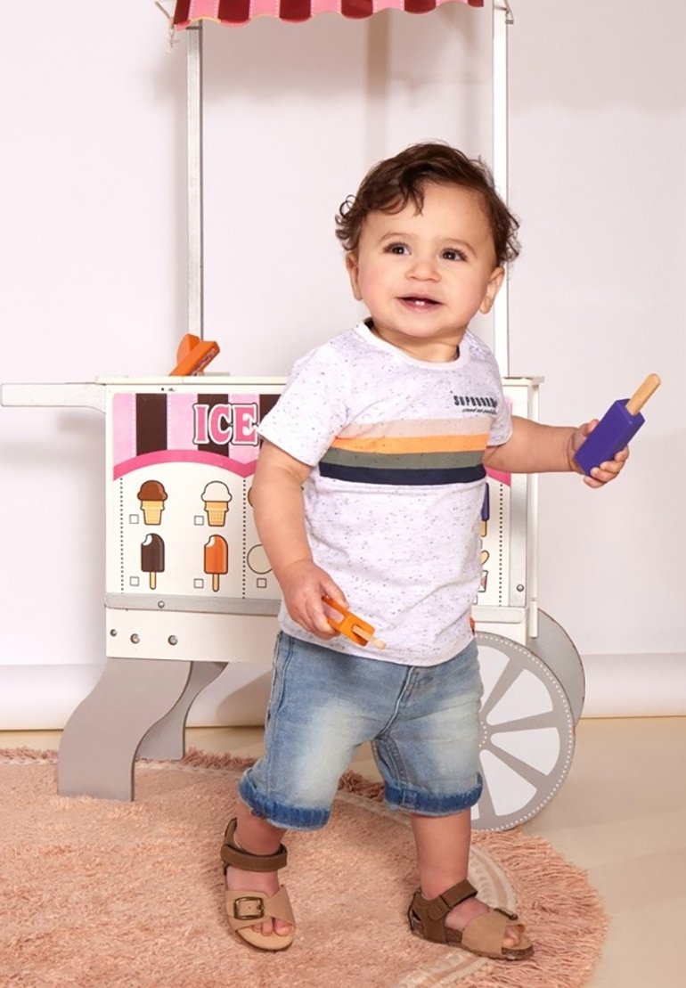 Peuter in een gespikkeld wit T-shirt met horizontale strepen, denim shorts en bruine sandaaltjes, die met speelgoedijsjes speelt. IJsjeskar op de achtergrond.