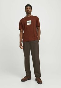 Brun T-shirt med et hvidt og sort logo på brystet, parret med mørkebrune bukser og brune sko. Simpelt design, korte ærmer.
