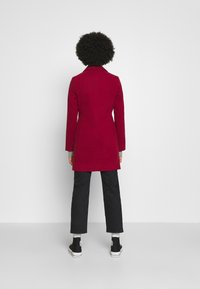 Rød frakke med skræddersyet design, der har en notch-krave og rygsøm. Kombineret med sorte bukser og højhælede sneakers.