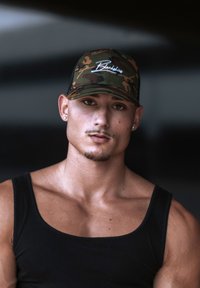 Kamerás baseball sapka "Blackskies" logóval, fekete ujjatlan felső, kidolgozott izmok, világos bőr és rövid haj, sötét, elmosódott háttér előtt.