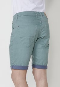 Shorts en coton verts avec un ourlet roulé, ornés de poignets bleus contrastants, de poches arrière avec un détail de couture et d'une étiquette sur la taille.
