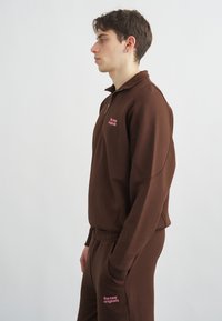 Jauns vīrietis, ģērbies brūnā sporta tērpā ar rozā uzrakstu "the new originals", stāv profilā pret vienkāršu baltu fonu.