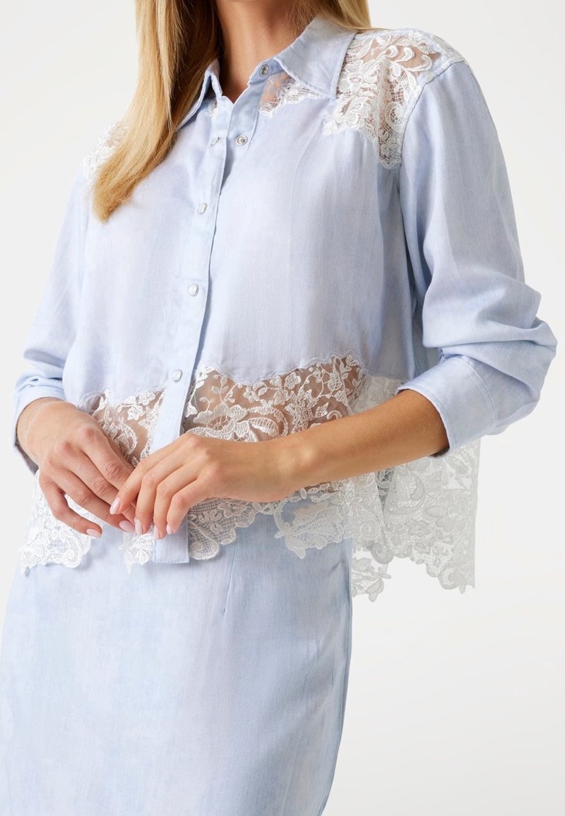 Jasnoniebieska koszula z długim rękawem, z koronkową wstawką na końcu. Zapięcie na guziki, miękka tkanina i przezroczyste koronkowe detale w kwiatowy wzór.