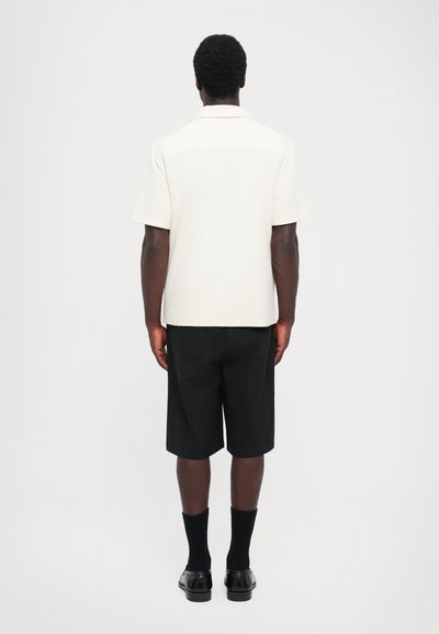 Kurzärmliges, cremefarbiges Hemd mit Knöpfen und strukturiertem Stoff, kombiniert mit schwarzen Shorts und schwarzen Socken, von hinten betrachtet.