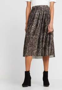 Mujer que lleva una falda midi plisada con un patrón de serpiente marrón y negro y botas de tobillo de ante negro con tacones cuadrados.