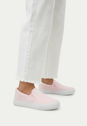 Person iført hvide korte bukser og lyserøde slip-on sneakers med hvide såler på en ensfarvet lys baggrund.