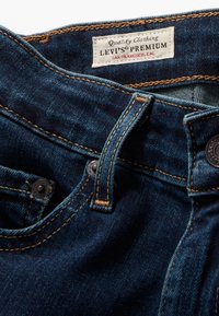 Tmavě modré džínové kalhoty s hnědým prošíváním, kovovými knoflíky a štítkem s nápisem "Levi's Premium" na bílém textilním rámci.