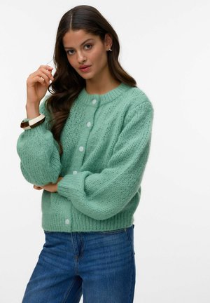 Kobieta ubrana w jasnografitowy sweter z grubym splotem i białymi guzikami oraz niebieskie dżinsy, pozująca z jedną ręką przy twarzy, a drugą skrzyżowaną.