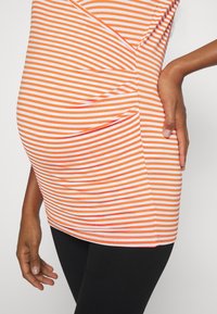 Top listrado em laranja e branco, com um design envolvente e pregas sobre a barriga grávida, combinado com leggings pretas.