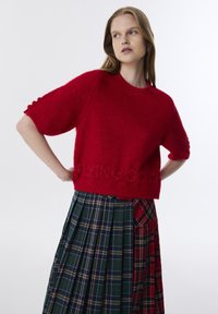 Roter gestrickter Pullover mit kurzen Ärmeln und Rundhalsausschnitt, mit eingeprägtem Text "FLYING SO" am Saum. Kariert plissierter Rock in mehreren Farben.