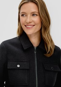 Czarna kurtka typu zip-up wykonana z teksturowanej tkaniny, z wyrazistym kołnierzem i dwoma przednimi kieszeniami z zapięciem na napy.