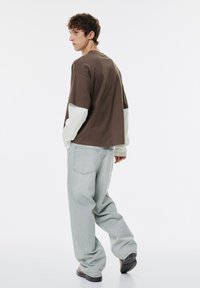 Brązowa, oversize'owa koszulka nałożona na jasnoróżową bluzkę z długim rękawem, w połączeniu z jasnoszarymi spodniami typu wide-leg. Czarne obuwie dopełnia stylizację.