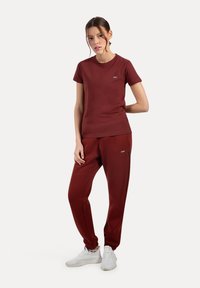 T-shirt color bordeaux e pantaloni joggers coordinati, entrambi realizzati in tessuto misto cotone. La T-shirt presenta un logo sul petto; i joggers includono tasche laterali.
