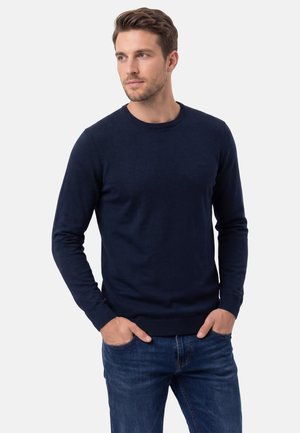 Man met kort haar, gekleed in een donkerblauwe trui en een blauwe spijkerbroek, met handen in de zakken, kijkt naar opzij tegen een witte achtergrond.