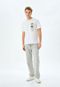 Camiseta blanca de algodón con un gráfico central que presenta árboles y paisajes en escala de grises. Combinada con jeans gris claro y zapatillas blancas.