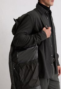 Zwarte donsjas met een glanzende, gladde afwerking, voorzien van een taillekoord, gedragen over een donkergeruit overhemd en een coltrui.