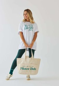 Biała oversize koszulka z zielonym tekstem graficznym, zielone legginsy i beżowa torba na ramię z zielonym napisem. Modelka trzyma torbę, nosząc trampki.