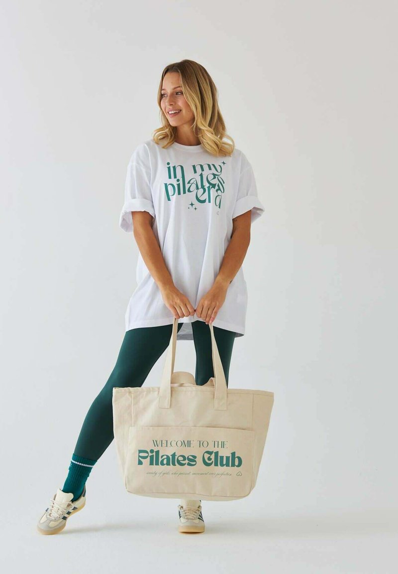 Biała oversize koszulka z zielonym tekstem graficznym, zielone legginsy i beżowa torba na ramię z zielonym napisem. Modelka trzyma torbę, nosząc trampki.