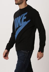 Černá mikina s modrým logem Nike, vyrobená z měkkého materiálu. Má dlouhé rukávy, klasický výstřih a ribbované zakončení. Nosená s tmavými džíny.