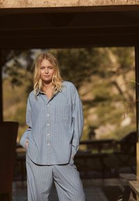 Modrý a bílý pruhovaný loungewear set vyrobený z měkké látky, který obsahuje košili na knoflíky s náprsní kapsou a volné kalhoty.