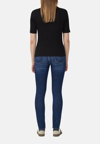 Haut noir à manches courtes associé à un jean slim-fit bleu. Le jean présente des poches arrière avec des accents beiges et une texture lisse. Chaussures décontractées visibles.