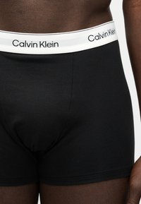 Torso masculino con bóxers Calvin Klein negros, cintura blanca con logotipo, mostrando detalles frontales y laterales.