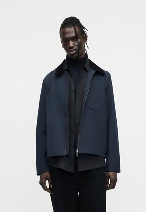 Άνδρας με πλεξούδες, φορώντας στενή μπλε ζακέτα με φερμουάρ πάνω από μαύρο πουκάμισο με κουμπιά και μαύρο παντελόνι, στέκεται μπροστά σε λευκό φόντο.