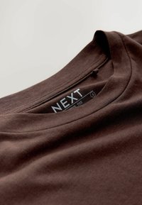 Tumši brūna kokvilnas T-krekls ar apaļu izgriezumu, uz kura ir birka ar uzrakstu "NEXT" un izmēru "S." Gluda tekstūra, vienkāršs dizains.
