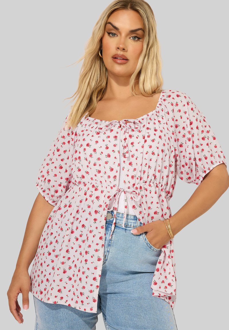 Mujer rubia con una blusa blanca con pequeño estampado floral rojo, mangas cortas y un lazo delantero, combinada con jeans azul claro y pulseras doradas.