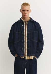 Temno modra teksas jakna z lahkotnim šivom, dvema sprednjima žepoma, nošena čez bež karo srajco, v kombinaciji z usklajenimi teksas hlačami.