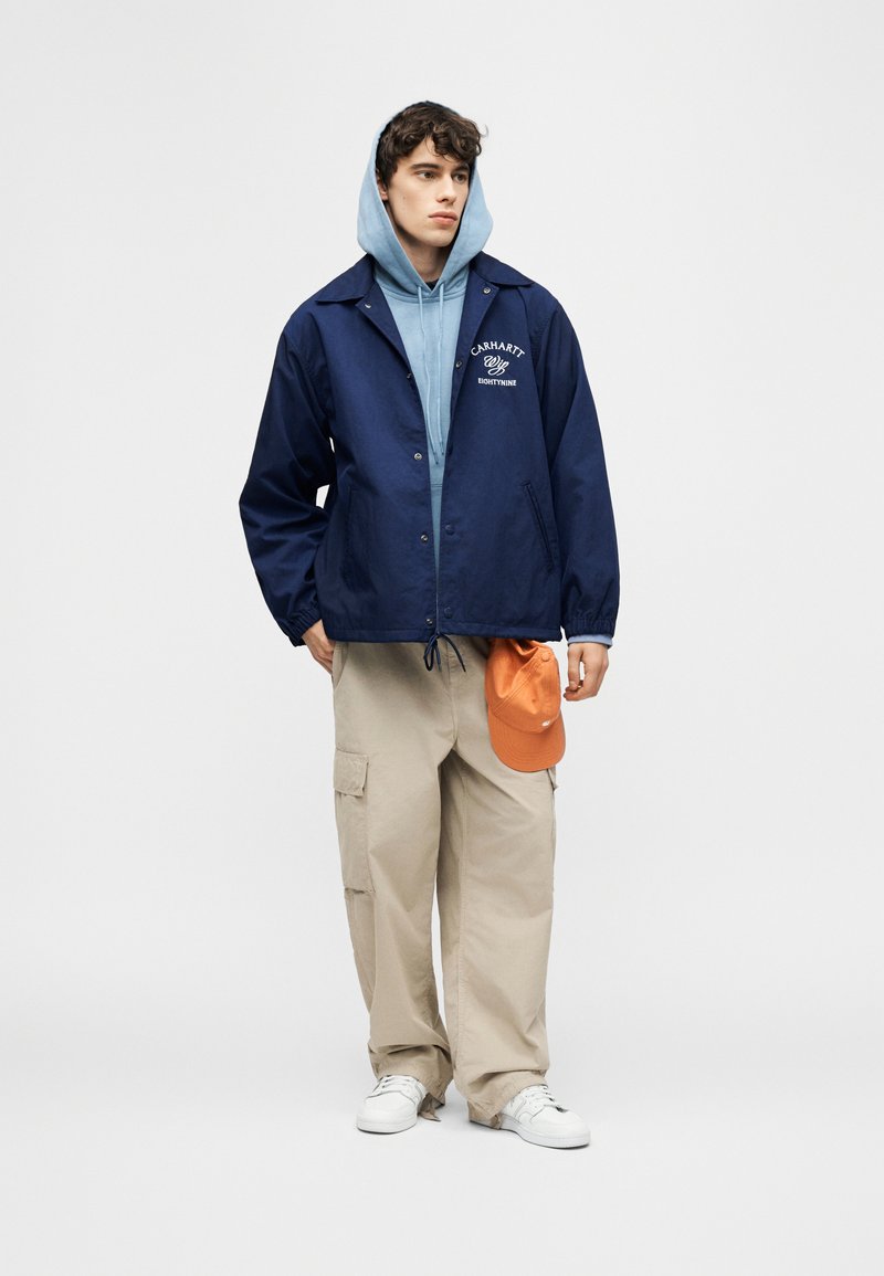 Giovane uomo con felpa con cappuccio blu, giacca blu scuro con scritte, pantaloni cargo beige, sneakers bianche, che tiene un cappellino arancione contro uno sfondo semplice.