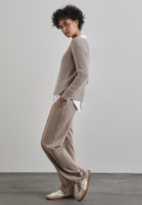 Ljust beige tröja med texturerad stickning, bärs över en vit skjorta. Tanbyxor med fickor, ihop med vita och tan sneakers. Stående pose.