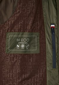 Jas interieur met bruine stof met het label "HECHTER," een groene patch met de tekst "H-ECO duurzame stof," en een ritszak met een driekleurige tab.