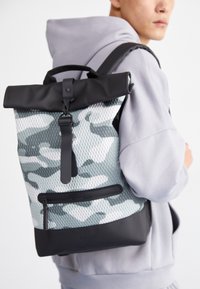 Zaino con motivo camouflage dotato di chiusura a rotolo, caratterizzato da dettagli neri, tessuto in rete testurizzata e una tasca con zip sul fronte.