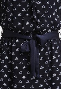 Mörkblå klänning med vita triangulära mönster, med ett knytbart midjebälte. Tillverkad av ett lätt tyg med en slät yta.