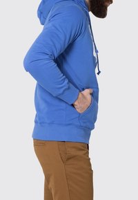 Sudadera azul con bolsillo canguro, capucha con cordón, puños y dobladillo acanalados. Llevada con pantalones marrón claro, mostrando una vista lateral.