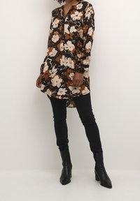 Femme portant une tunique fleurie marron, noire et beige, avec un pantalon skinny noir et des bottines à talons noires, debout devant un fond uni.