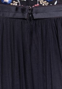 Jupe noire avec une fermeture éclair au dos et une ceinture, comprenant un tissu transparent plissé et des détails floraux brodés sur le bord supérieur.