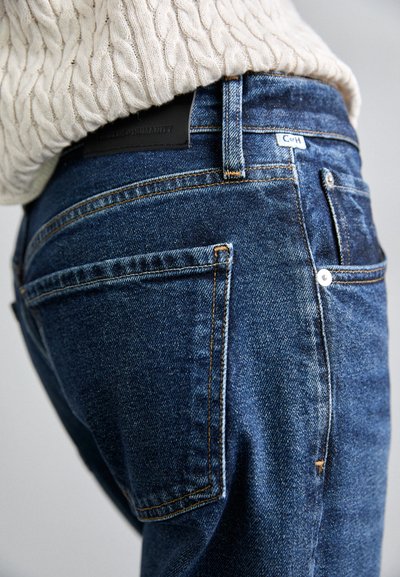 Donkerblauwe denim jeans met een aansluitend ontwerp, voorzien van oranje stiksels, twee achterzakken en een leren taillebanddetail.