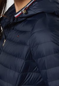 Marinblå puffjacka med horisontellt kviltat mönster, försedd med huva, dragkedjestängning och kontrasterande randiga detaljer vid halsen.