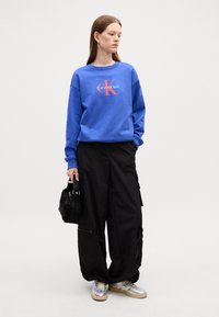 Felpa blu con grafica rossa "Calvin Klein", abbinata a pantaloni cargo neri e sneakers argentate. Il modello tiene una borsa nera.