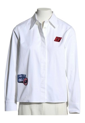 Chemise blanche à manches longues avec col et boutons, décorée de patchs rectangulaires rouges et bleus sur la poitrine et la partie basse avant.