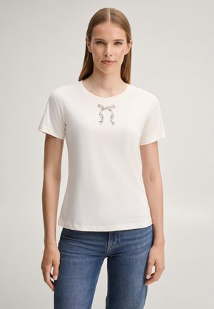 Vrouw staand, gekleed in een effen wit T-shirt met korte mouwen en een klein decoratief strikje, en blauwe jeans tegen een neutrale achtergrond.