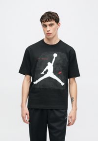 Μαύρο βαμβακερό μπλουζάκι με λευκό γραφικό Air Jordan με μοτίβο πλέγματος, κοντά μανίκια και στρογγυλή λαιμόκοψη. Συνοδεύεται από μαύρο παντελόνι.