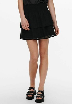 Kobieta nosząca krótką czarną spódnicę warstwową i czarne platformy na sandałach z trzema paskami z klamrami, stojąca na białym tle.