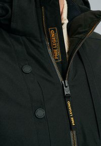 Giacca nera con interno trapuntato, dettagli arancioni della cerniera e bottoni tondi. Presenta il marchio "PME Legend" sull'etichetta e sul tessuto.