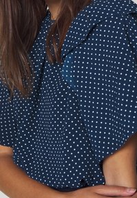 Marinblus med vita prickar, med en volangkrage och korta ärmar, tillverkad av mjukt tyg med en slät yta.