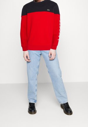 Homme portant un sweat-shirt Lacoste à blocs de couleur rouge et noir, un jean bleu clair et des bottes noires à lacets, se tenant devant un fond blanc.